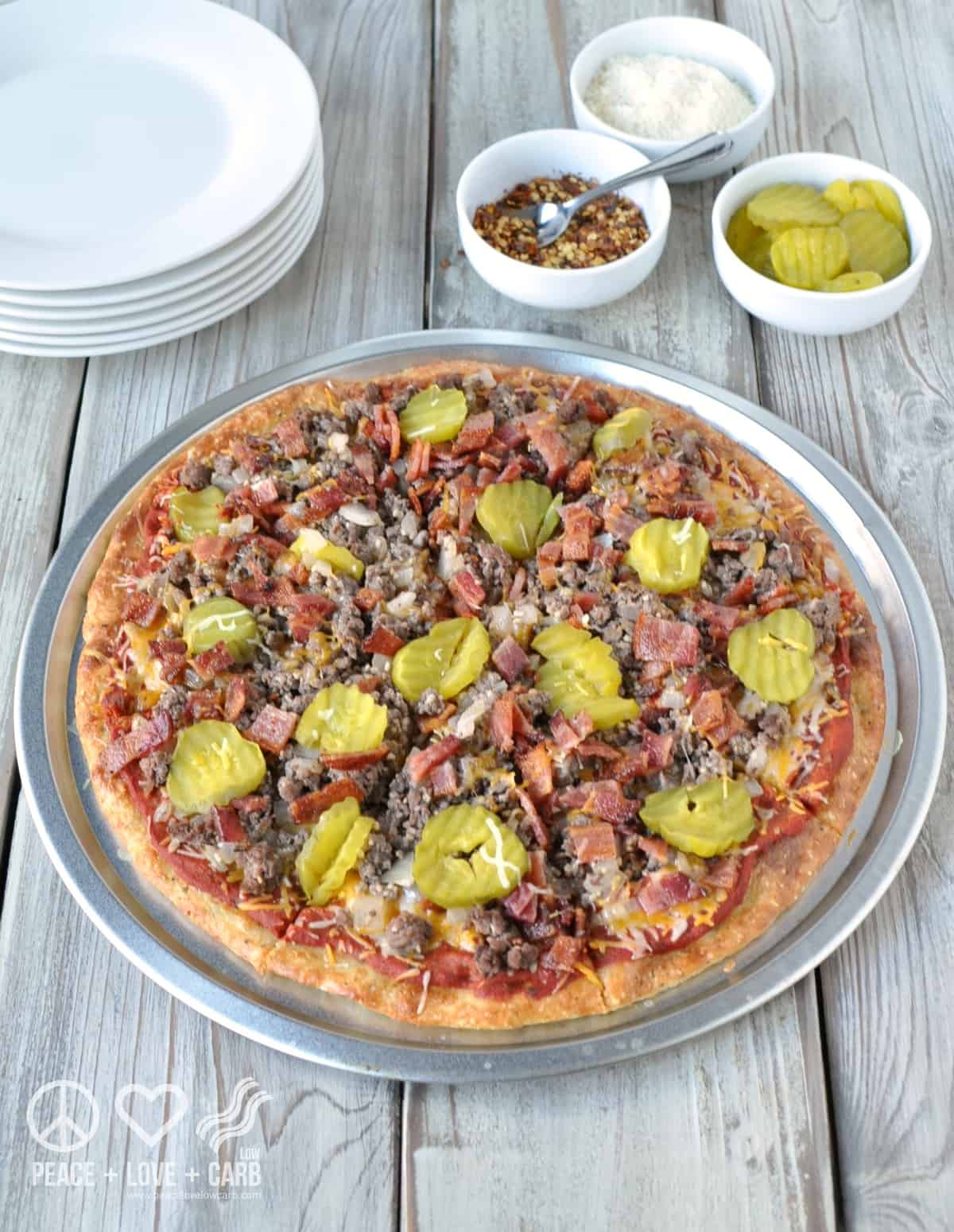 A bacon cheeseburger pizza sits on a metal pizza charger on a white wooden table. In the upper left corner of the frame sits a stack of white round dishes. To the upper right is 3 white pinch bowls filled with red pepper flakes, shredded cheese, and pickles.