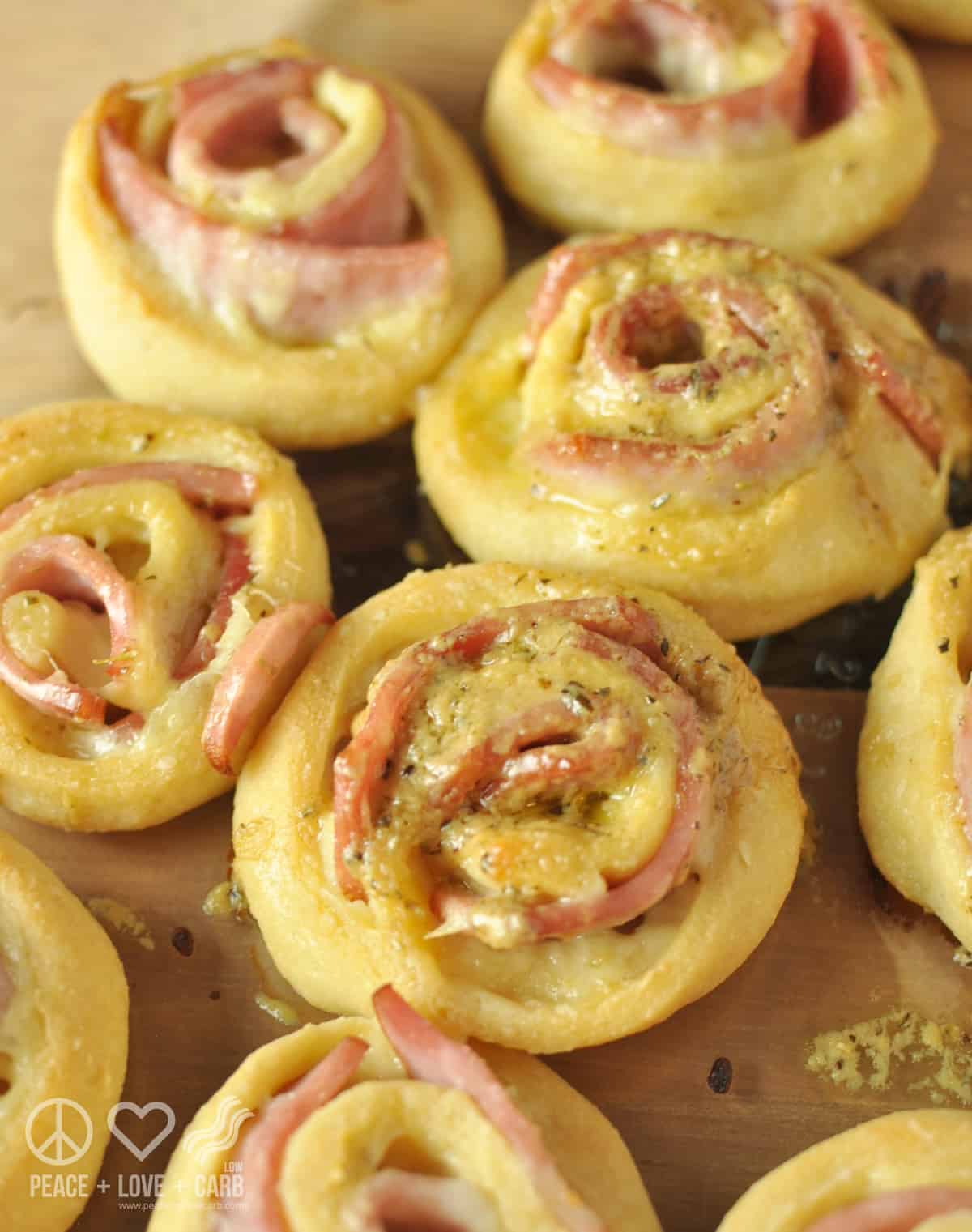 Individual baked hot ham and cheese roll ups, coated with honey dijon butter glaze and sprinkled with herbs, sitting on a wooden cutting board.