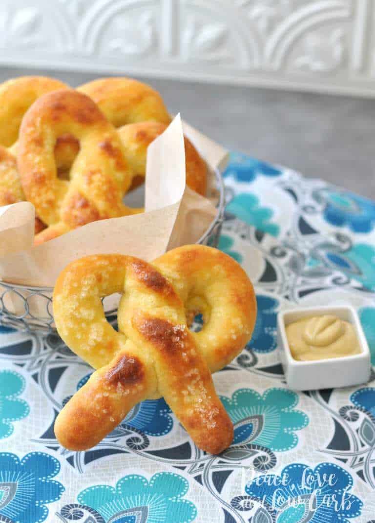 On a gray countertop with a white patterned backsplash, a metal basked with parchment paper loaded with keto soft pretzels sits to the left side of the frame on top of an aqua, gray, black, and blue tablecloth. At the center of the photo sits a golden brown keto soft pretzel with a small white square sauce dish full of mustard.