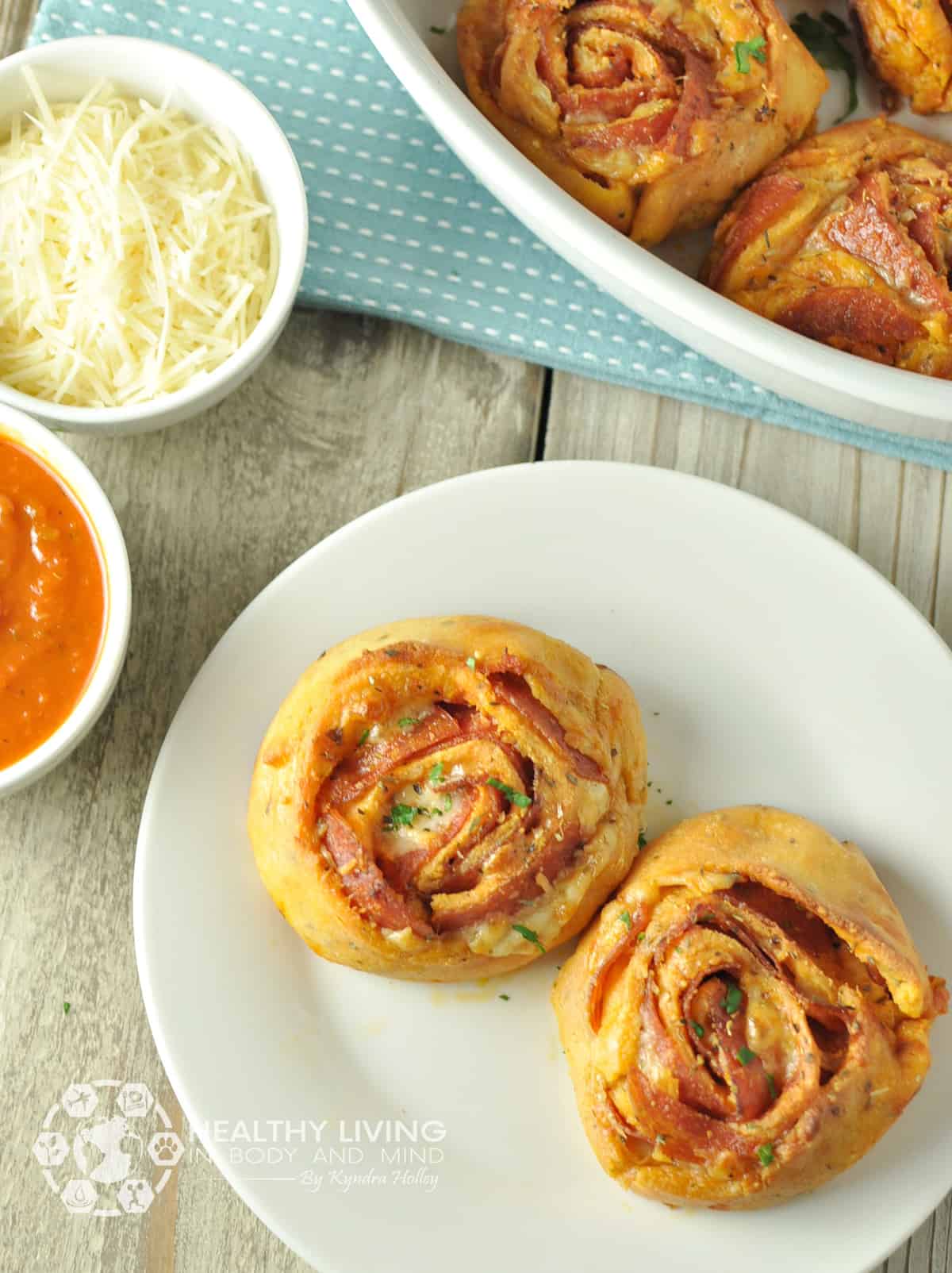 Two pepperoni pizza pinwheels sit on a white plate on a gray wooden table. To the left is a white round sauce dish filled with pizza sauce, and another above it with shredded cheese. In the upper right corner is a bowl of more pepperoni pizza pinwheels.