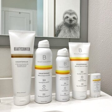 Beautycounter sunscreen lined up on a bathroom vanity: Countersun After Sun Cooling Gel, Countersun Mineral Sunscreen Mist SPF 30, Countersun Mineral Sunscreen Lotion SPF 30, and Countersun Mineral Sunscreen Stick SPF 30. Above the bathroom vanity is a mirror that reflects back a framed photo of a sloth.