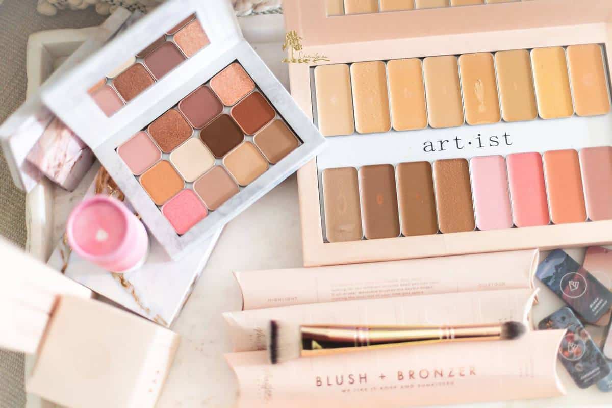 Overhead shot of a flat lay of Maskcara Makeup products: a white eyeshadow palette filled with warm tones; a large peach foundation palette filled with a row of highlight colors and a row of contour and lip and cheek colors; plus a double ended brush, a few foundation tins, and packaging.