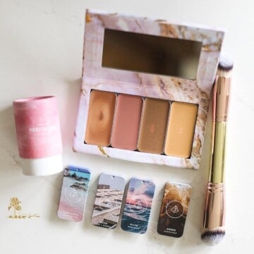Overhead shot of a flat lay of Maskcara Makeup products: a pink marble palette with 4 large tins of product, plus the 4 tin lids for each product below the palette; the package for a pink perfector sponge; and a double ended foundation brush, all sitting on a white background.