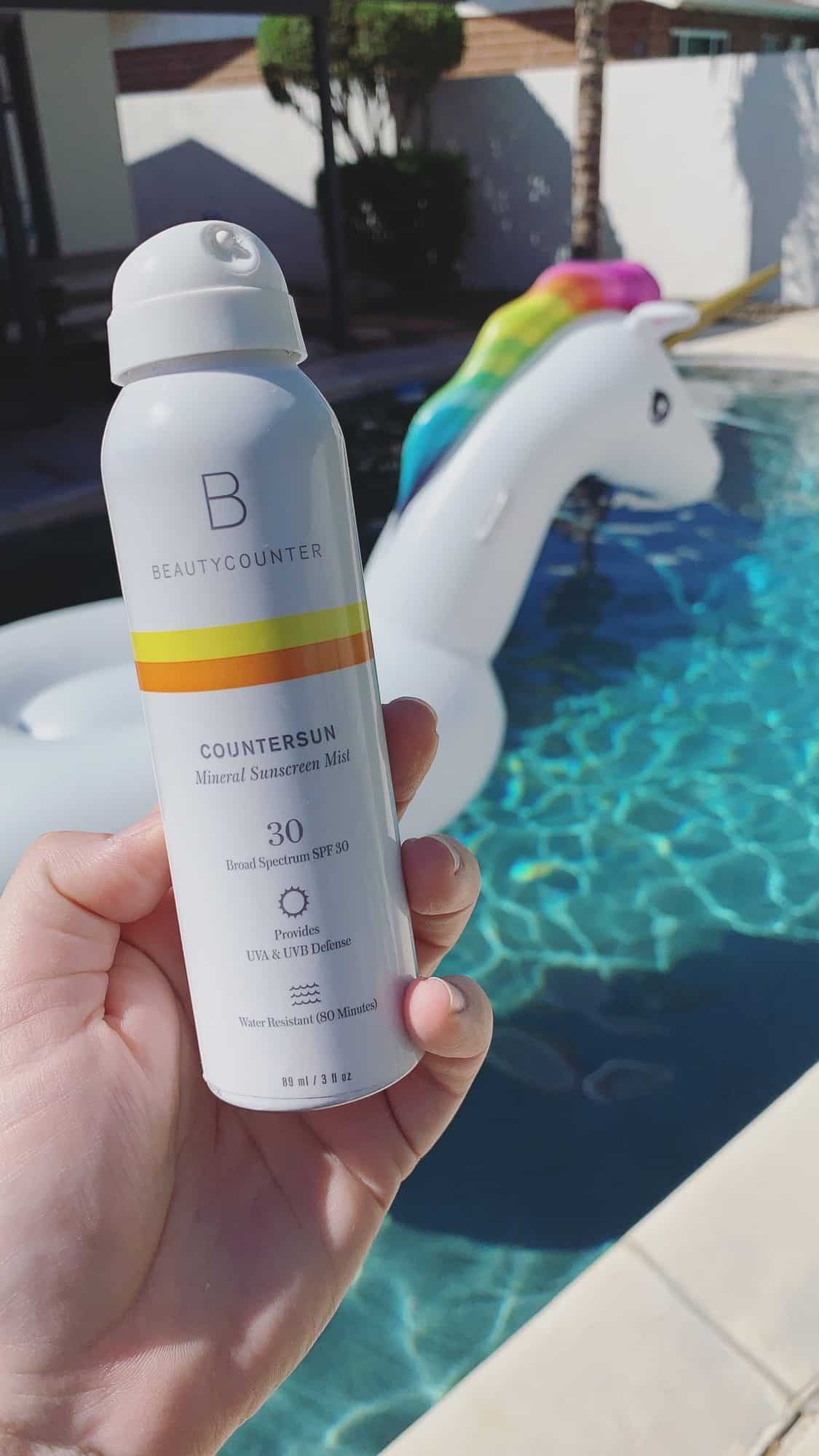 A hand holding a bottle of Countersun Mineral Sunscreen Spray in front of a pool with a large unicorn floaty in the background.