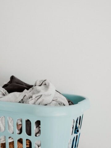 A pile of laundry in a light teal laundry basket sits against a white wall.