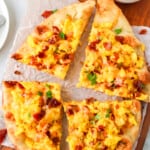 naan bread pizza on a wood cutting board, topped with scrambled eggs, cheese, and bacon