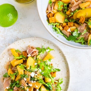 a white plate piled high with a fresh summer salad of prosciutto, peaches, melon, walnuts, and arugula