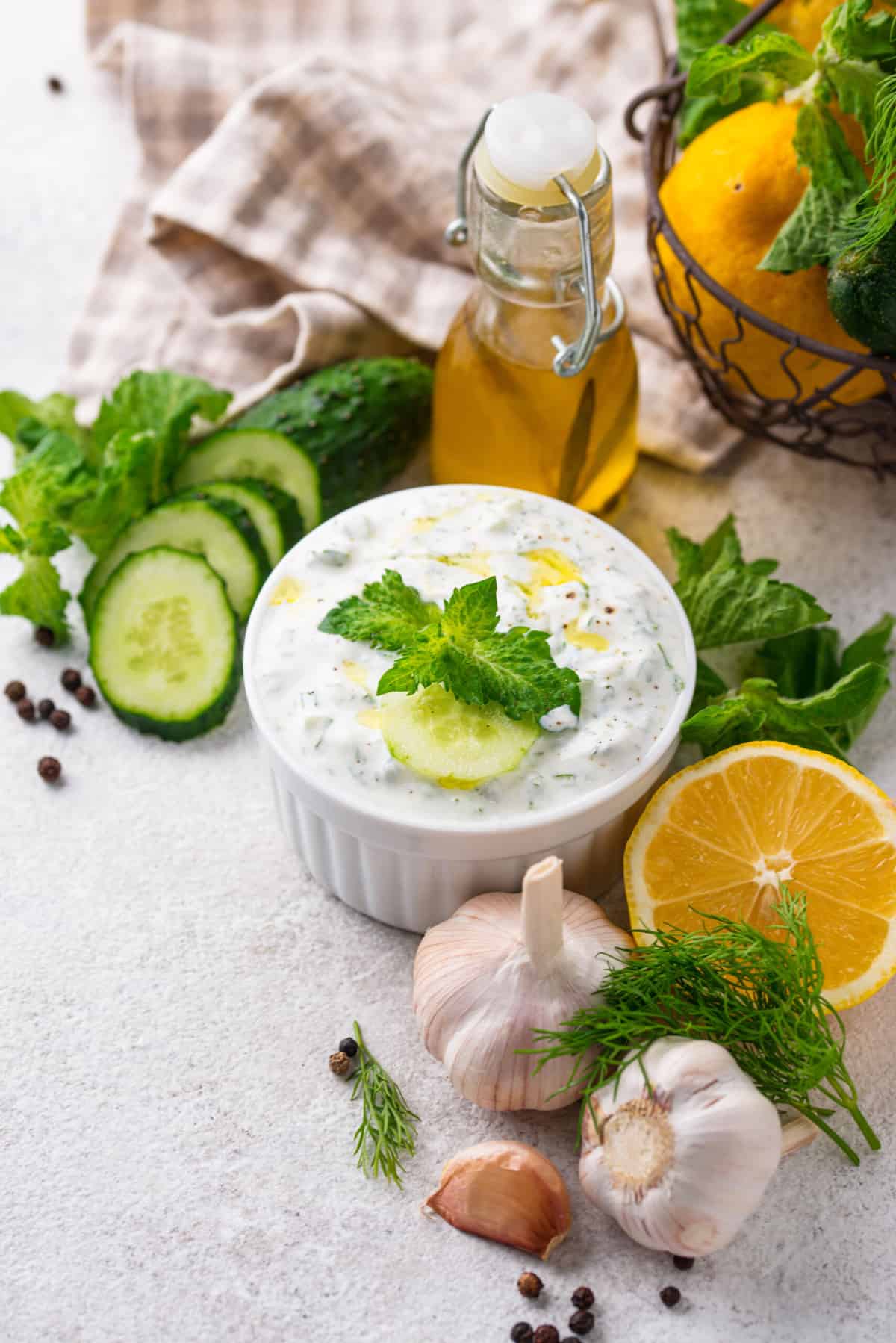 a white ramekin with greek cucumber sauce, surrounded by fresh herbs, lemons, garlic, and cucumbers