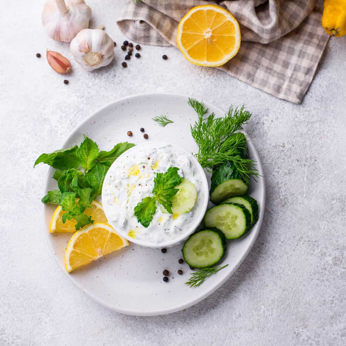 a white ramekin with greek cucumber sauce, surrounded by fresh herbs, lemons, garlic, and cucumbers