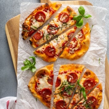 flatbread pizzas topped with pepperoni, cheese, and basil, on parchment paper, on a wood cutting board