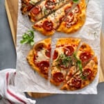 flatbread pizzas topped with pepperoni, cheese, and basil, on parchment paper, on a wood cutting board