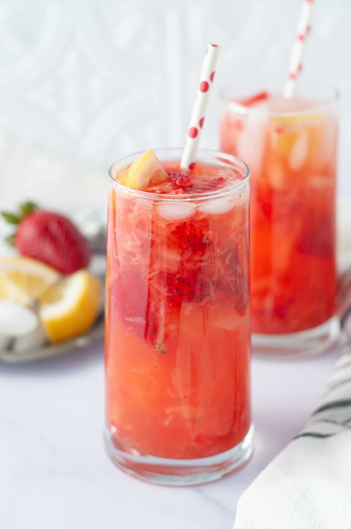 Tall classes with a strawberry lemonade cocktail in them. Red and white striped straw, garnished with strawberries and lemons