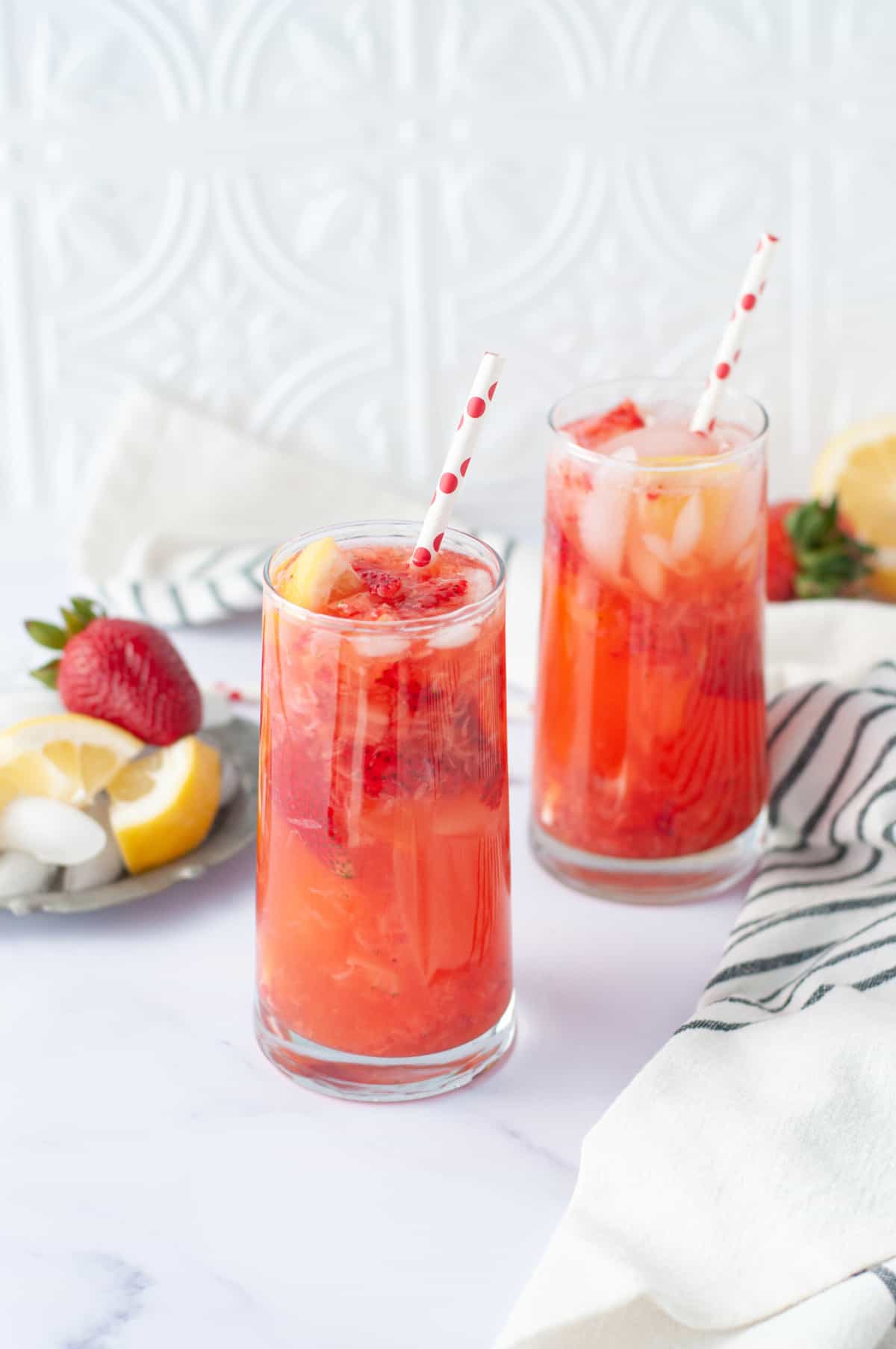 Tall glasses with a strawberry lemonade cocktail in them. Red and white striped straw, garnished with strawberries and lemons