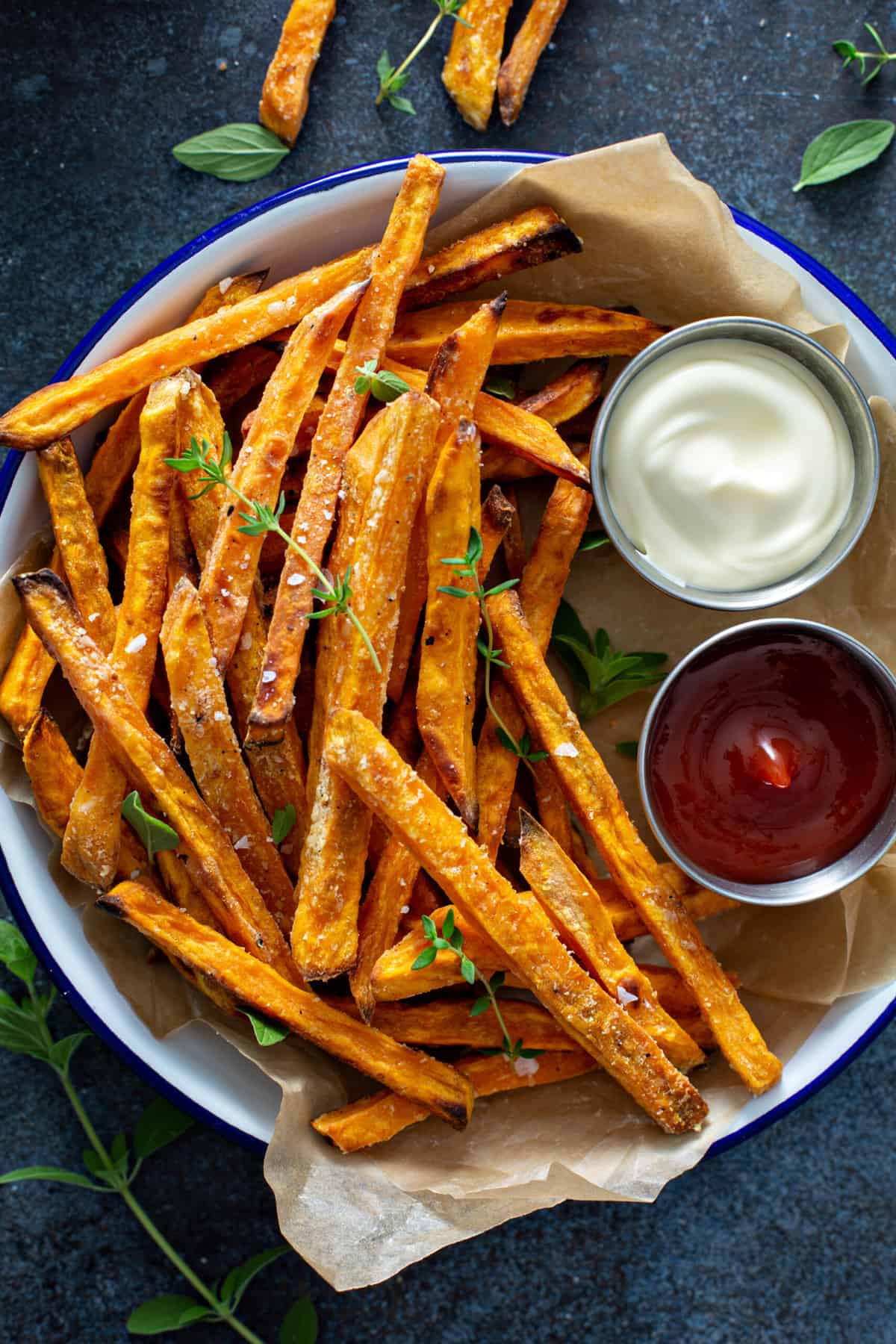 A serving bowl lined with parchment paper, filled with crispy air fryer sweet potato fries, sea salt and fresh herbs, served with ketchup and aioli.