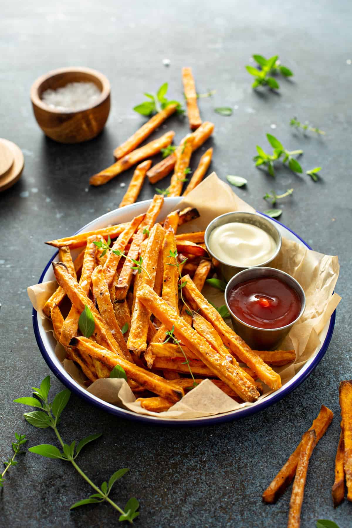 A serving bowl lined with parchment paper, filled with crispy air fryer sweet potato fries, sea salt and fresh herbs, served with ketchup and aioli.