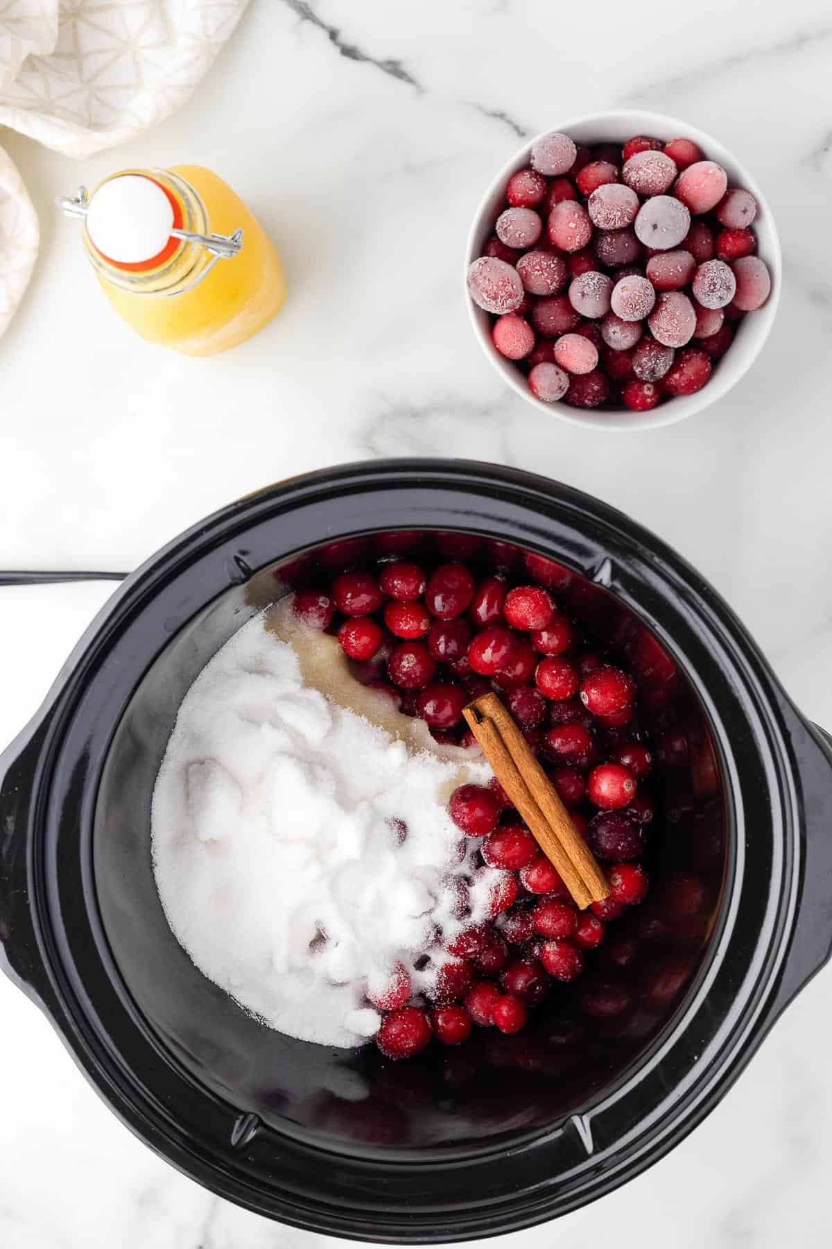 a slow cooker with cranberries, sugar, orange juice and a cinnamon stick