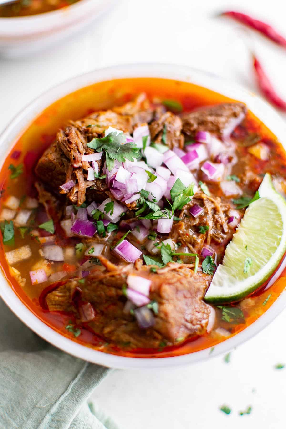 a bowl of beef birria Mexican stew, garnishes with red onion, cilantro and lime