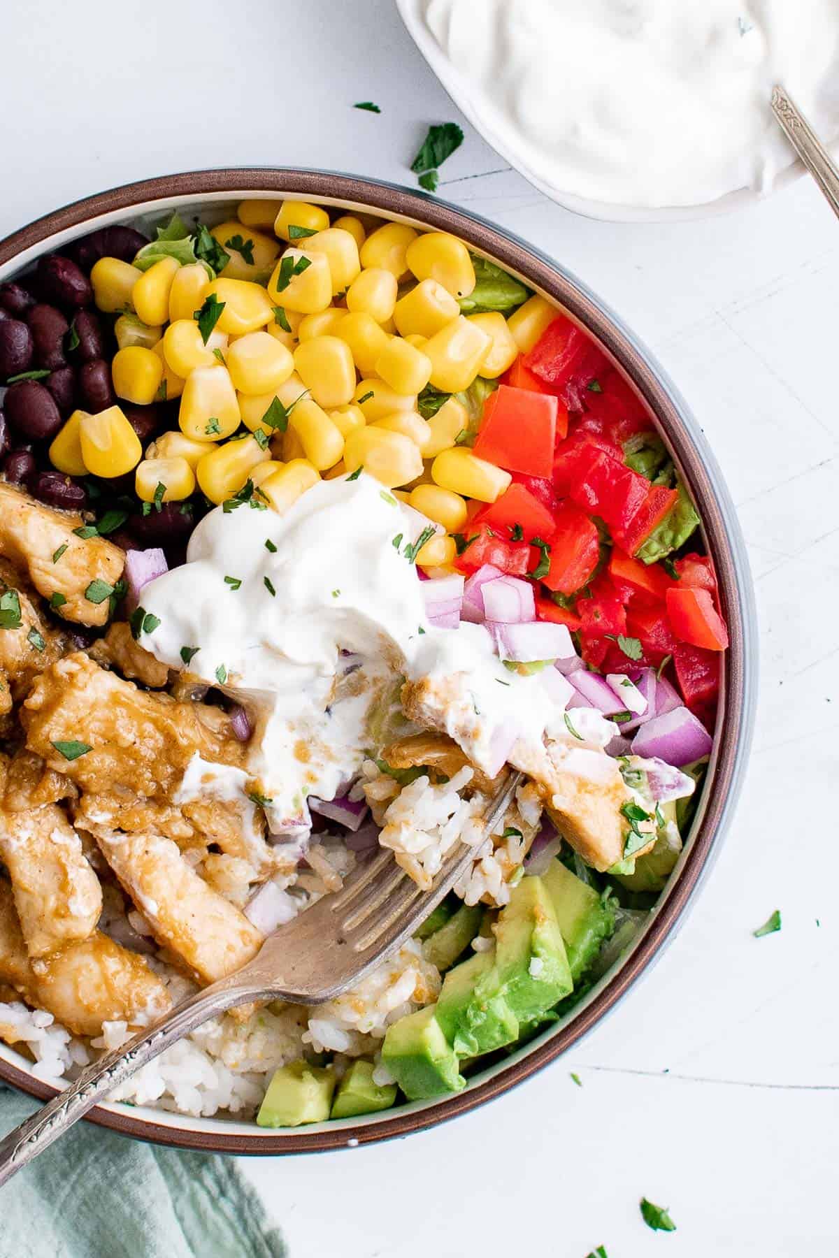 a burrito bowl, filled with chicken, onion, corn, beans, rice, avocado, and sour cream with a bite taken out of it and a fork