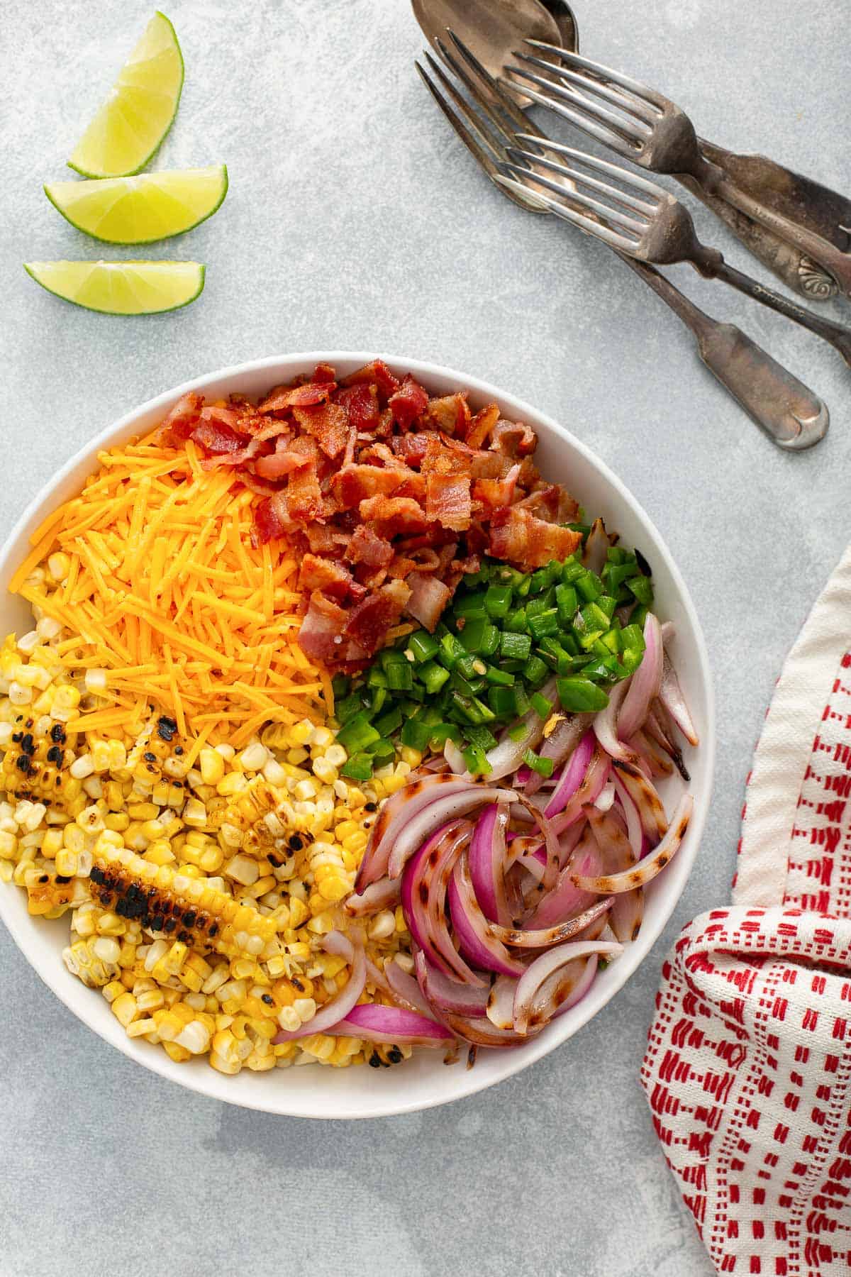 a white bowl with ingredients for grilled corn salad - corn, bacon, cheese, jalapeños, red onion, cheese, lime