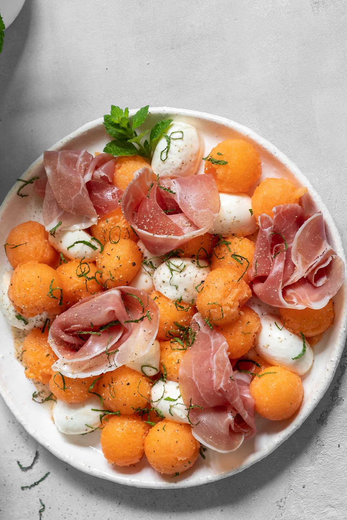 white plate with a salad made of thinly sliced prosciutto, cantaloupe balls, mozzarella cheese, mint and basil.