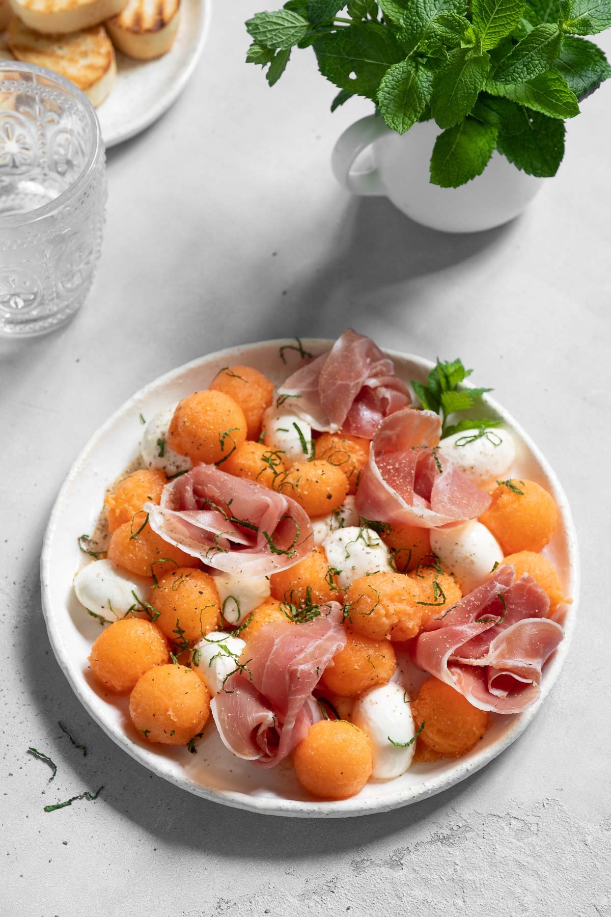 white plate with a salad made of thinly sliced prosciutto, cantaloupe balls, mozzarella cheese, mint and basil.