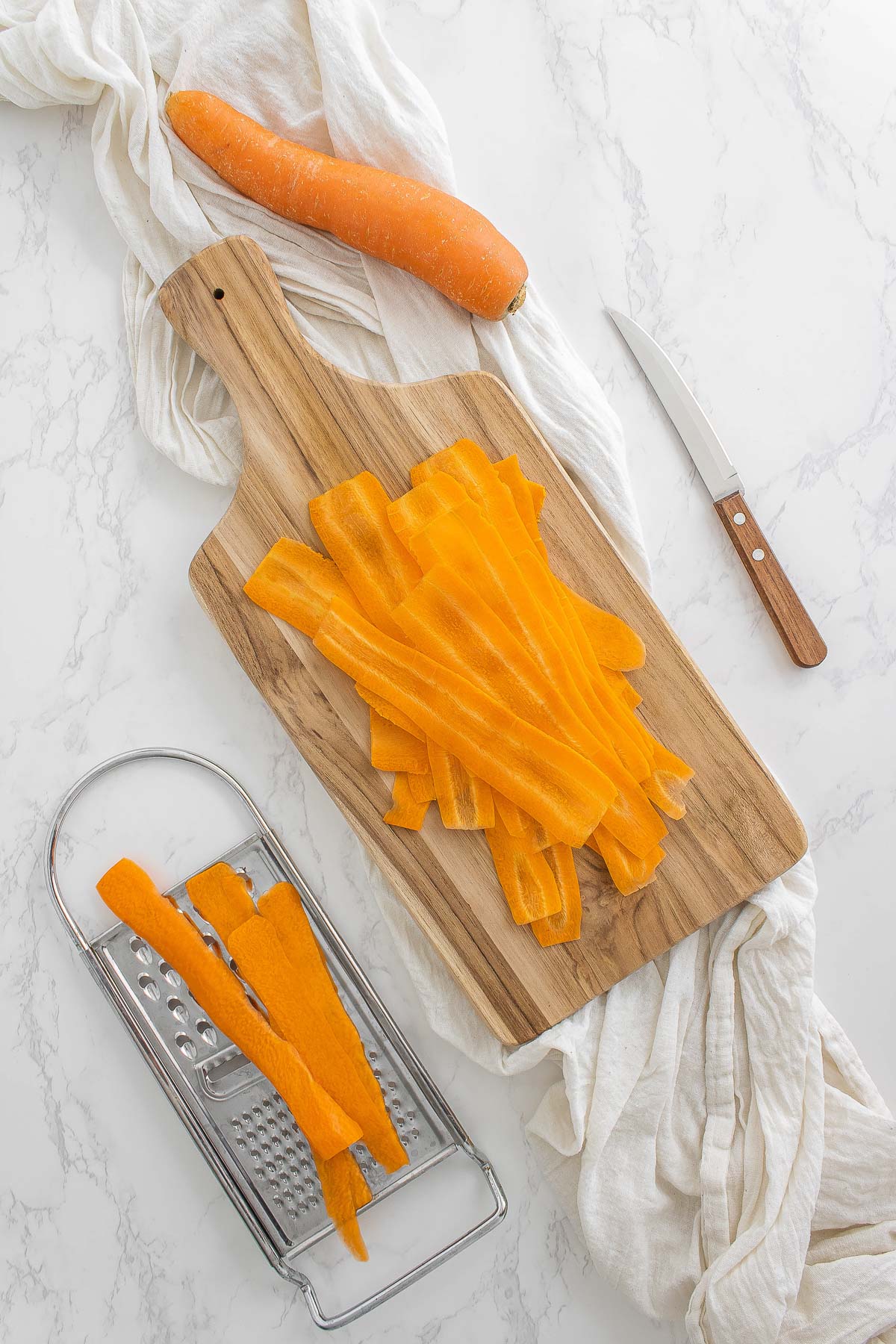 carrots being sliced on a grater with sliced on a wooden cutting board, with a knife.