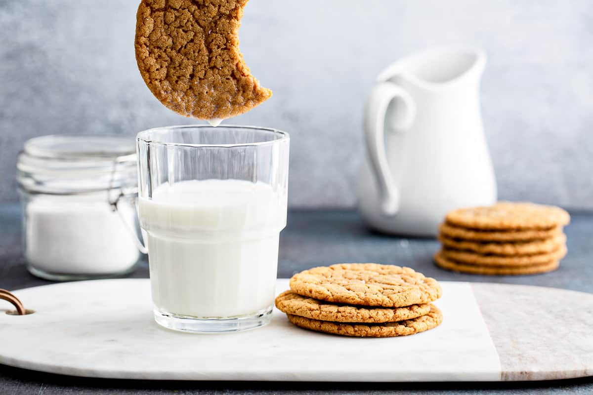 a stack of freshly backed gingersnap cookies on a white marble serving platter, with a glass of milk.