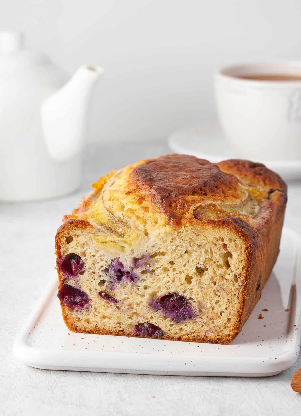 A white ceramic plate with a loaf of homemade banana bread with blueberries. There is a teapot and a mug of tea in the background.