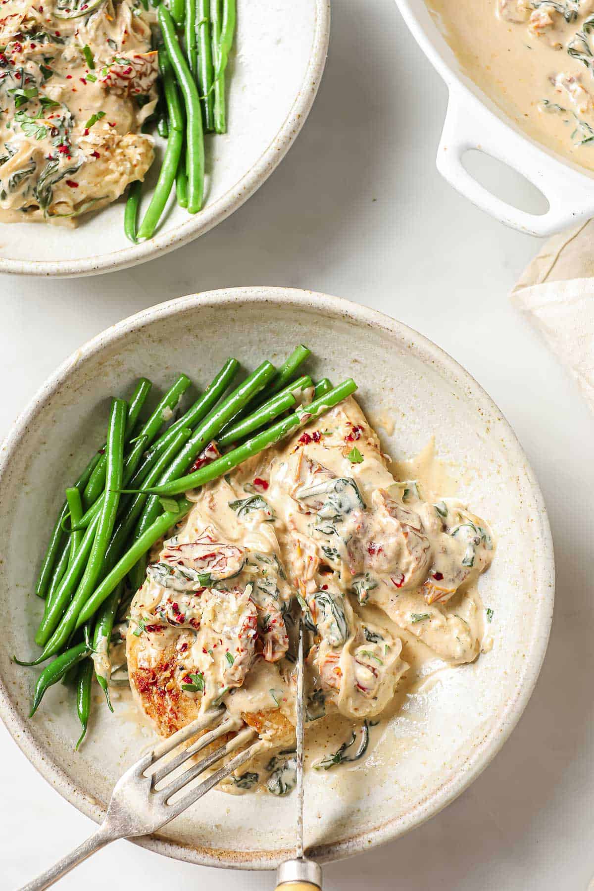 2 white bowls full of a creamy chicken dish with sun-dried tomatoes and spinach, served with a side of green beans.