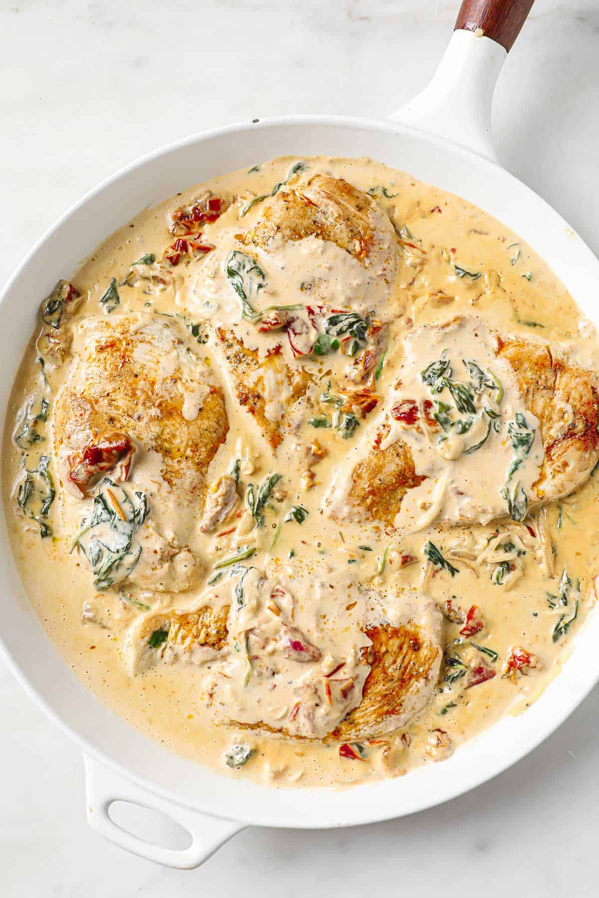 A white skillet filled with a chicken dish with a creamy tomato sauce, spinach and sun-dried tomatoes.