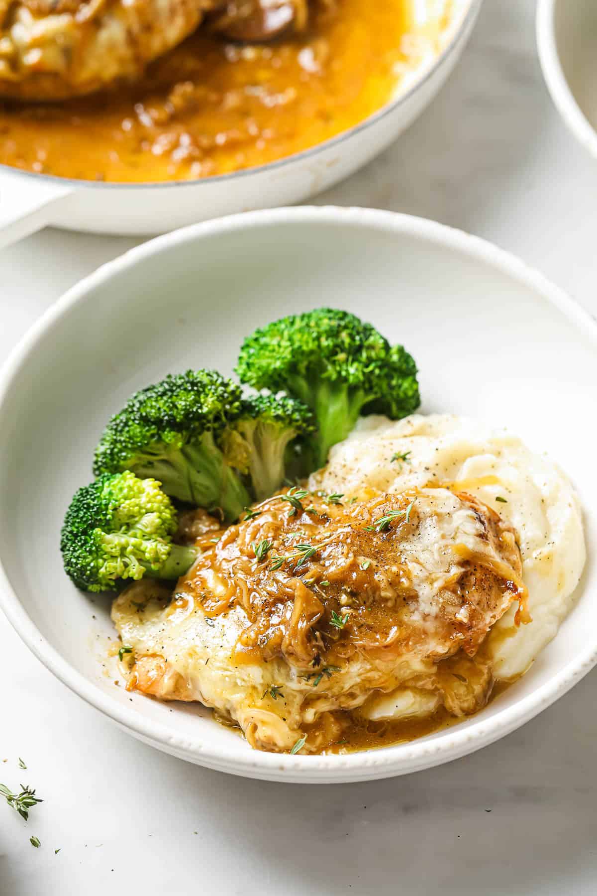 A white bowl with chicken and caramelized onions, mashed potatoes and roasted broccoli.