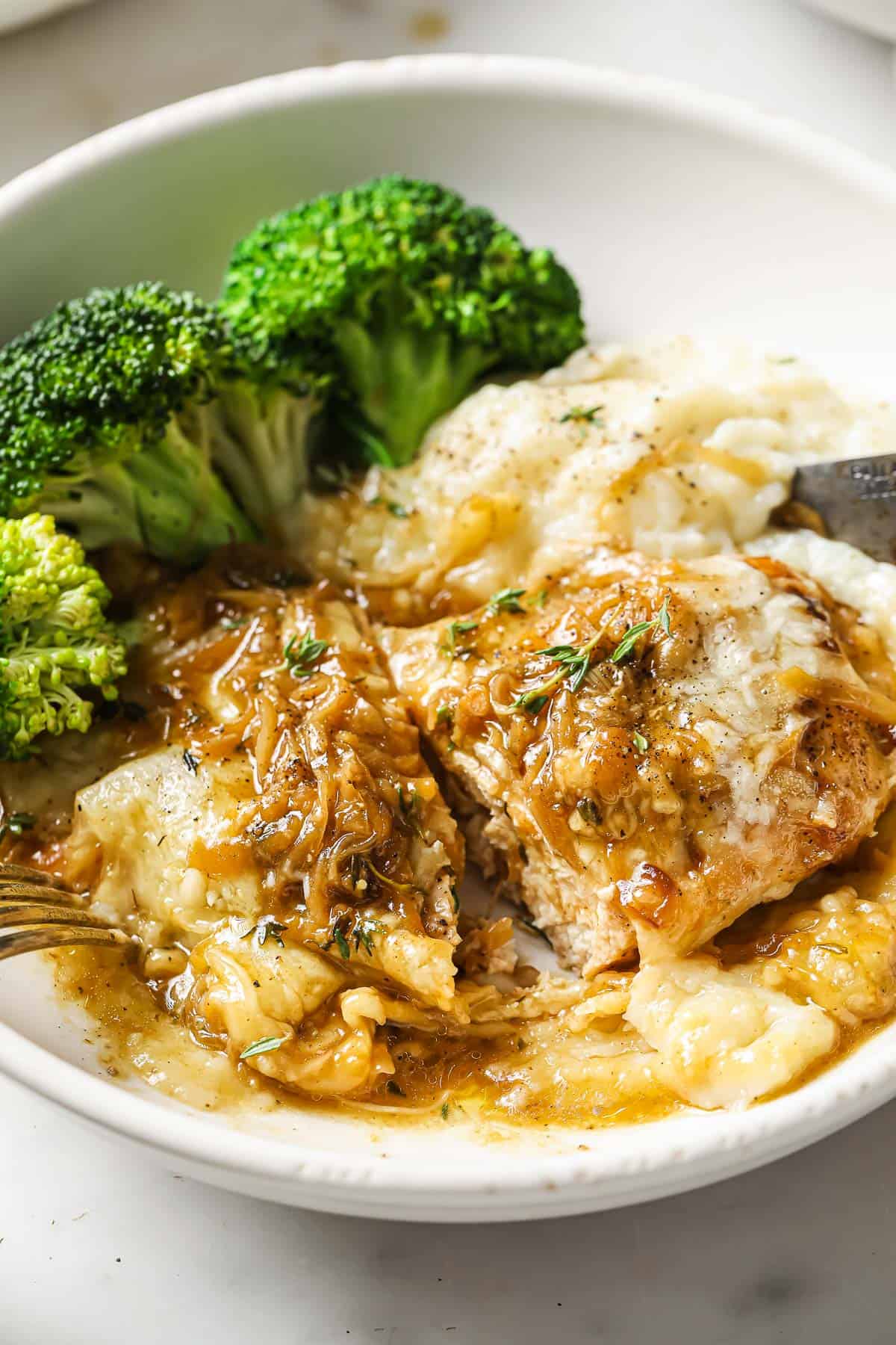 A white bowl with chicken and caramelized onions, mashed potatoes and roasted broccoli.