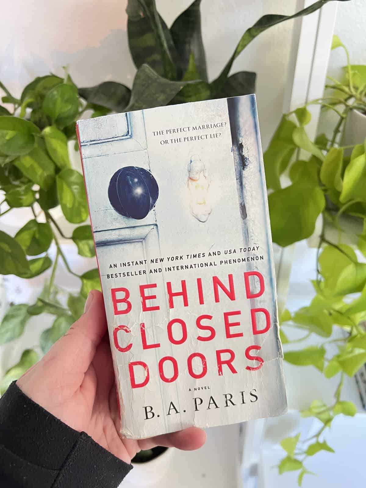 A paperback book of Behind Closed Doors by B.A Paris with a bunch of green plants behind it.