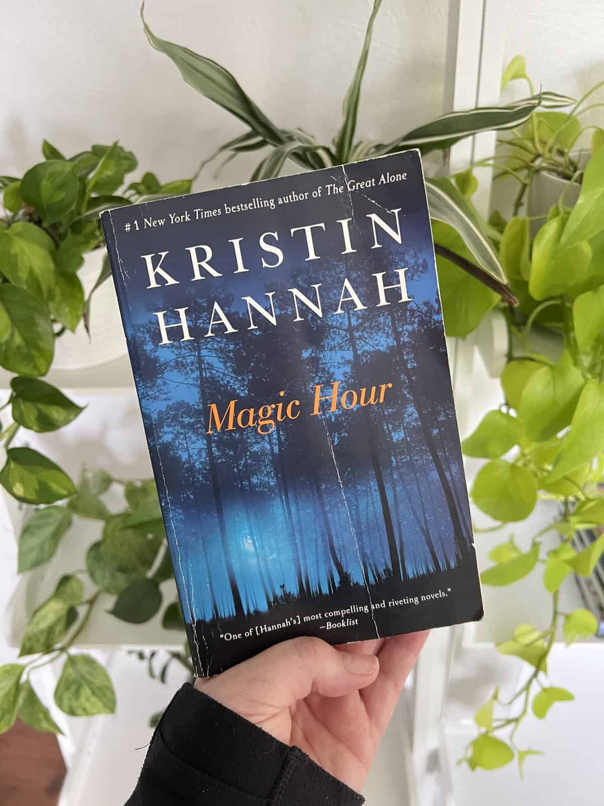 A paperback book copy of Magic Hour by Kristin Hannah with a bunch of green plants behind it.