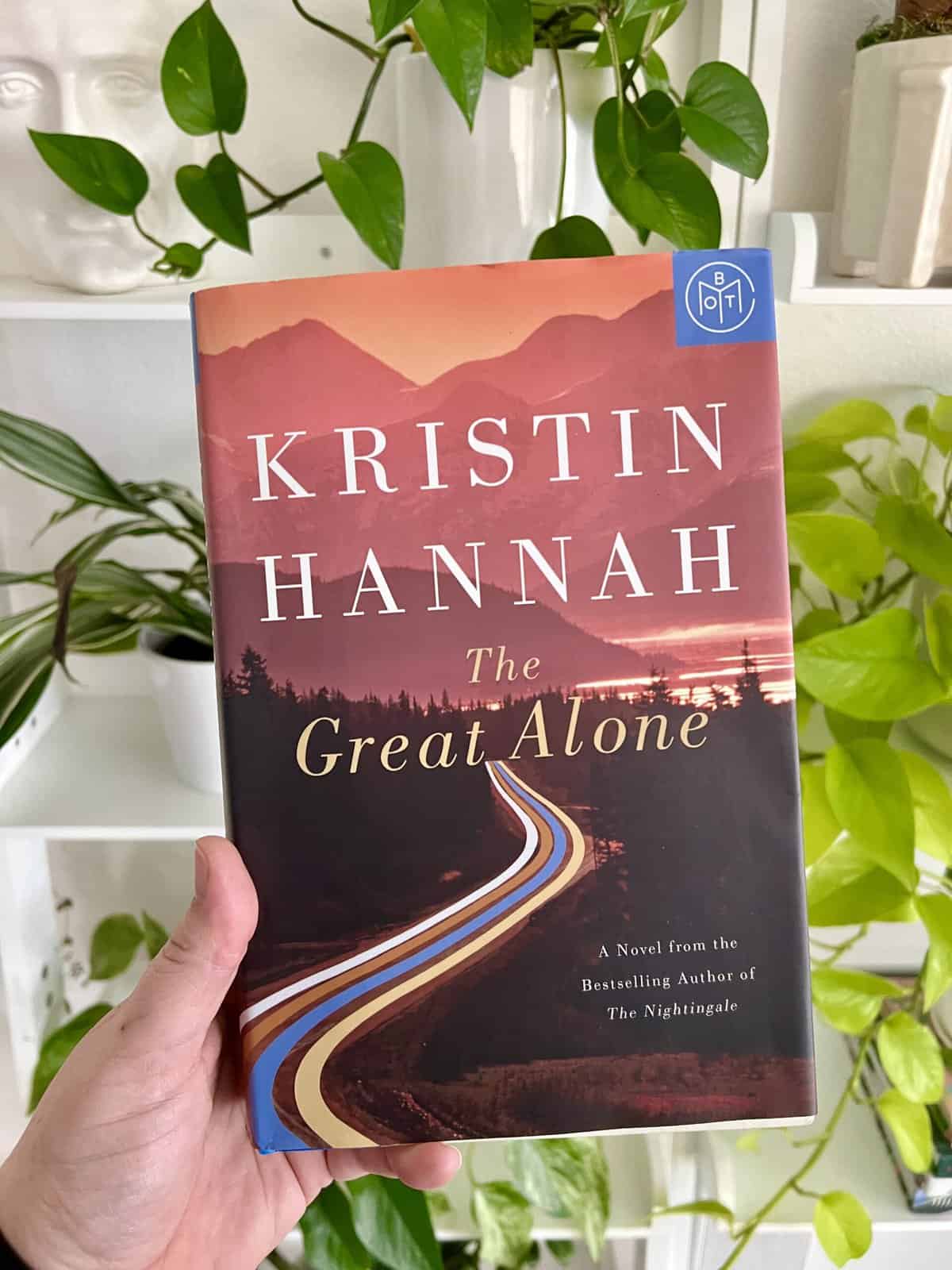 A hardcover book copy of The Great Alone by Kristin Hannah with a bunch of green plants behind it