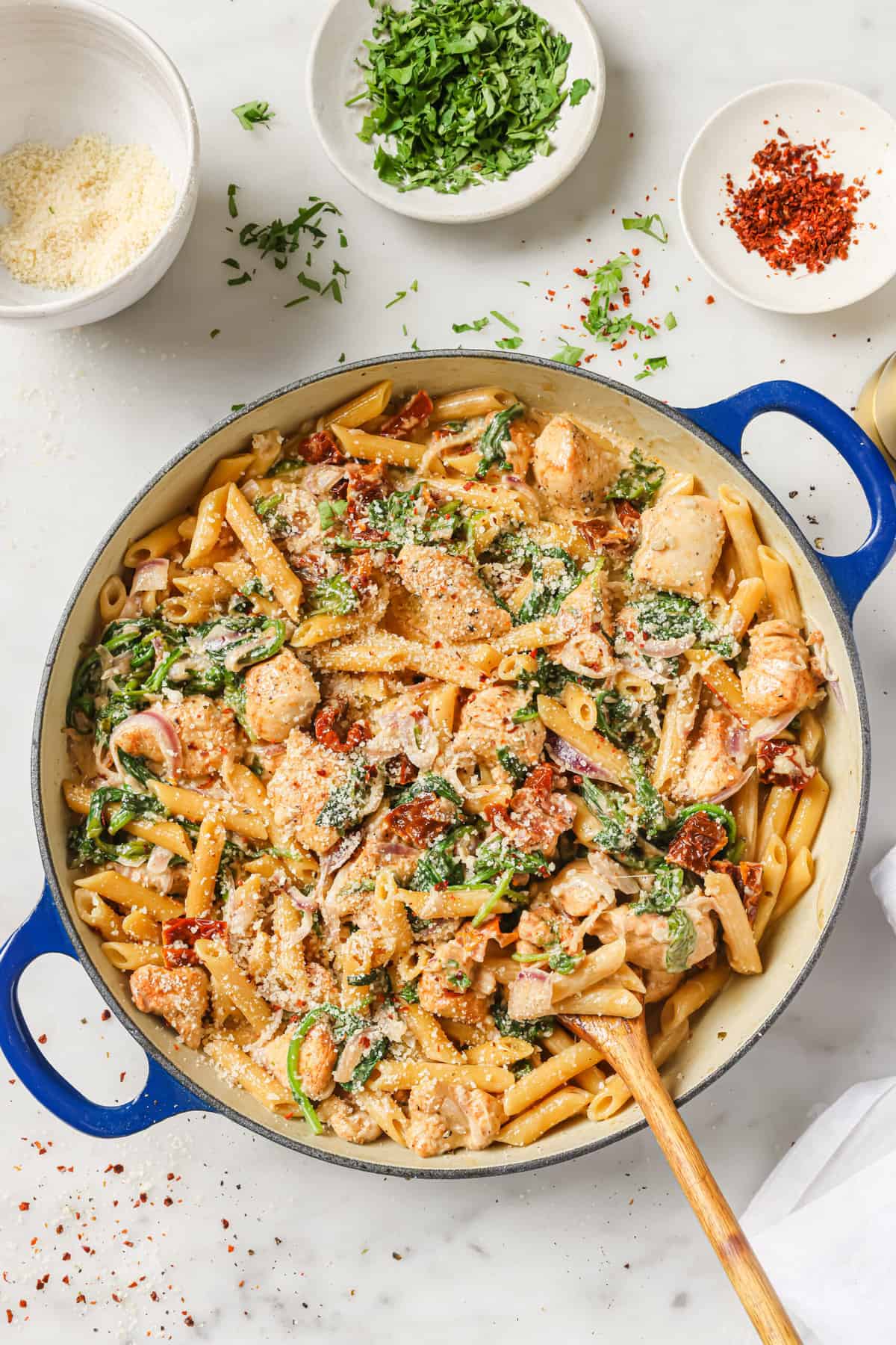 An enameled cast iron skillet filled with a pasta dish made with chicken, penne, sun-dried tomatoes, spinach, red onion and a rich creamy sauce