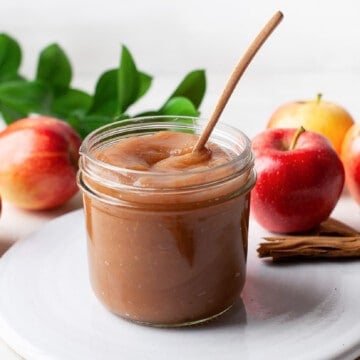 A jar of apple butter with a wooden spoon and apples and fresh herbs in the background.