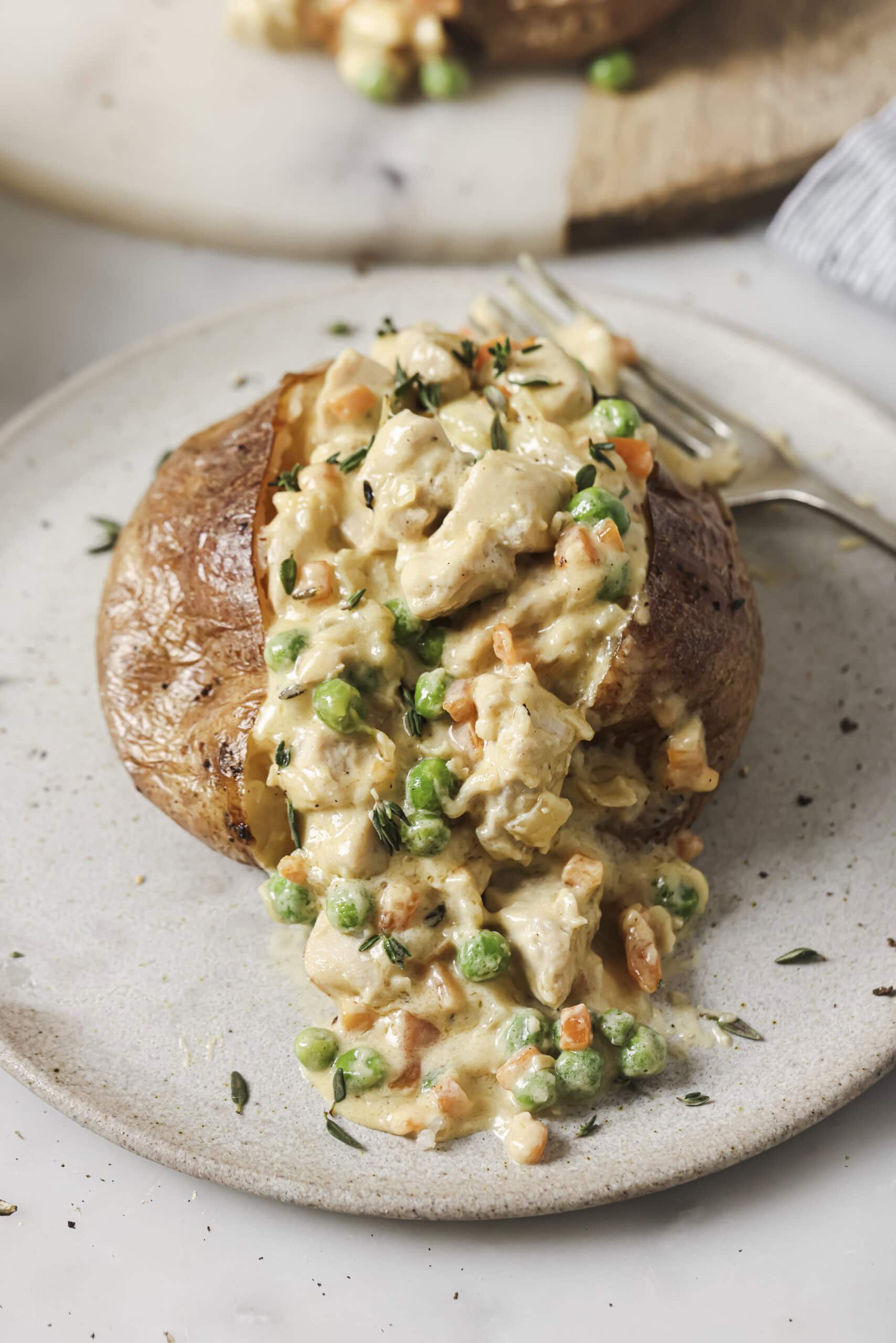 A close up shot of a baked potato on a ceramic plate, stuffed with a creamy gravy with chicken and vegetables.
